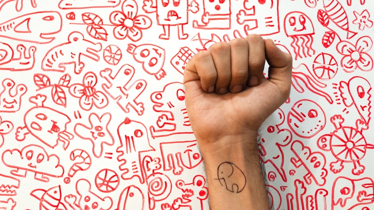 Hand with an elephant tattoo in front of a wall filled with artworks